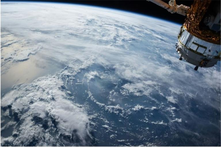 View of Earth from space with a satellite in the top right corner, showing clouds over a large body of water.