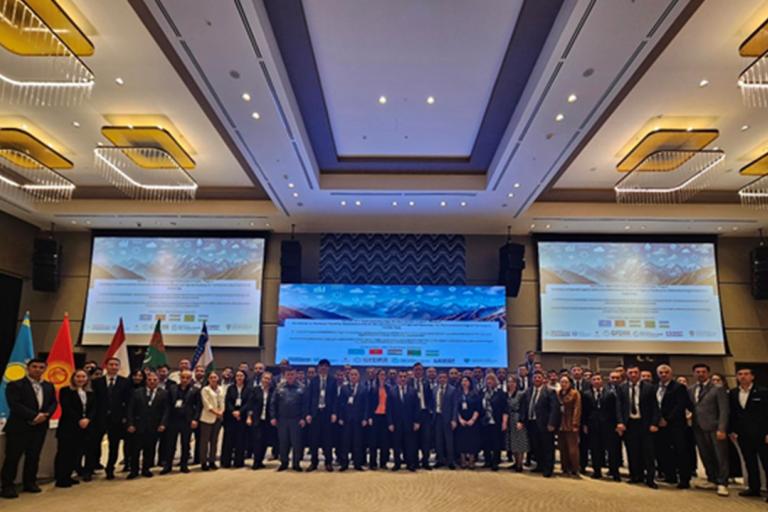 A group of people in formal attire posing in a large conference hall with multiple flags and two large screens in the background.