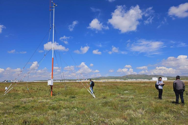 Two people stand beside a tall, slender weather station tower on a grassy field under a blue sky with white clouds.