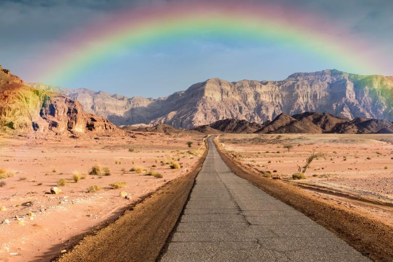 A straight road stretches through a rocky desert landscape with a rainbow arching above distant cliffs under a cloudy sky.