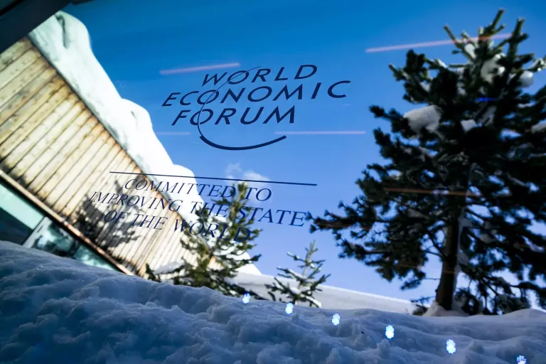 World Economic Forum sign on a glass panel, snow and trees reflected, with a snowy landscape and clear blue sky.
