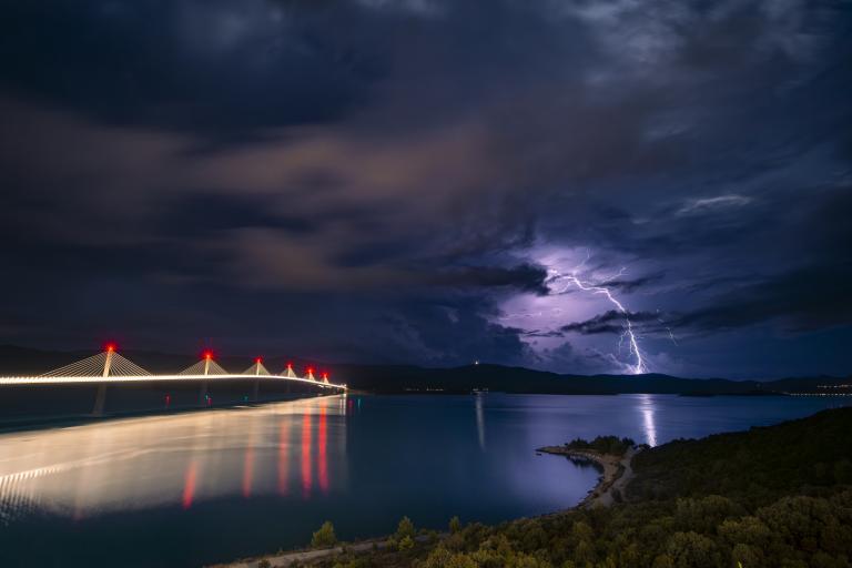 Lightning strikes over water near a long bridge with red lights under a stormy night sky.
