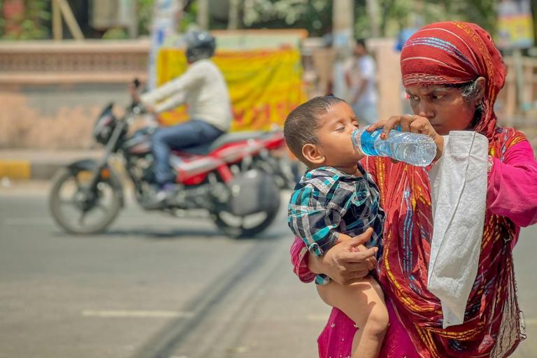 A woman in a colorful outfit gives water from a plastic bottle to a young child on a street with a motorbike passing by.