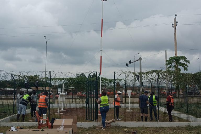 Workers in safety vests install a tall, red and white pole within a fenced area on a cloudy day. Nearby, boxes and wooden planks are on the ground.