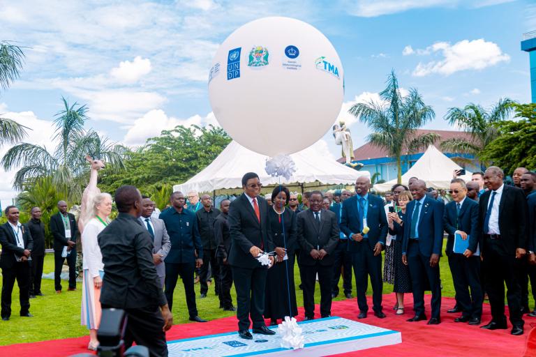 A group of people, including officials in suits, stand on a red carpet outdoors beneath a large white balloon with logos, surrounded by greenery and tents.