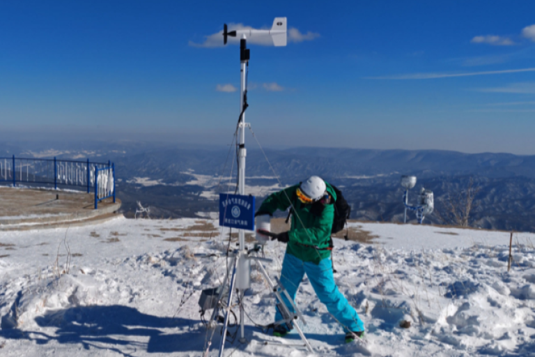 Person in winter gear adjusts a weather station on a snowy mountain summit under a clear blue sky.