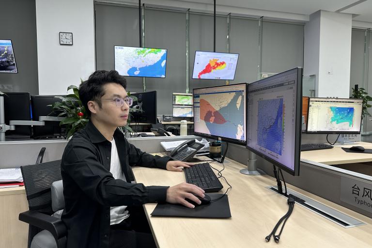 A person sits at a desk in an office with multiple monitors displaying weather maps and data.