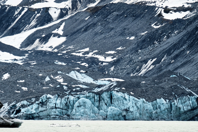A glacier with blue ice formations is surrounded by rocky, snow-dusted mountains, and a body of water is visible in the foreground.