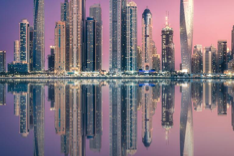 City skyline with tall, illuminated skyscrapers reflecting in a calm body of water during twilight. The sky is a gradient of pink and purple hues.
