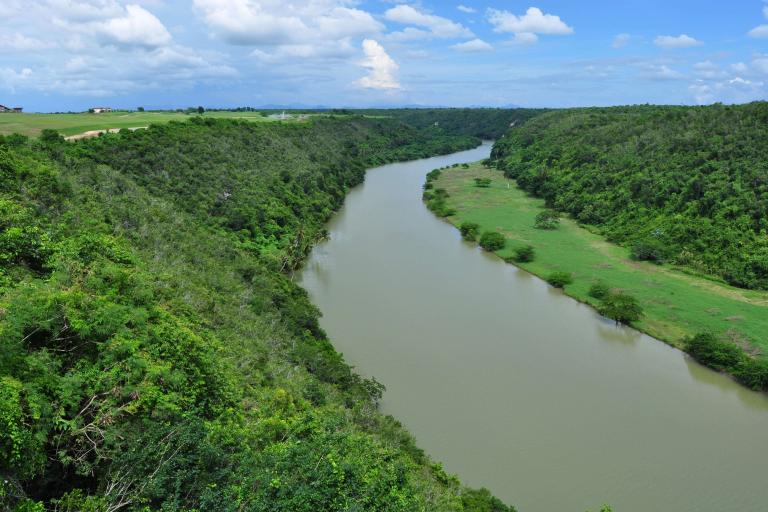 Aerial view of a wide river flowing through a lush, green valley, surrounded by dense forest and grassy slopes under a blue, cloud-dotted sky.