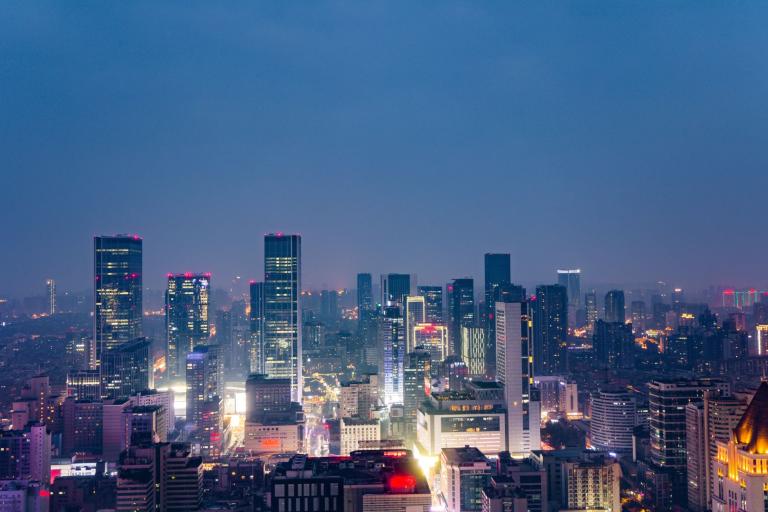Aerial view of a city skyline at night with illuminated skyscrapers and buildings under a deep blue sky.