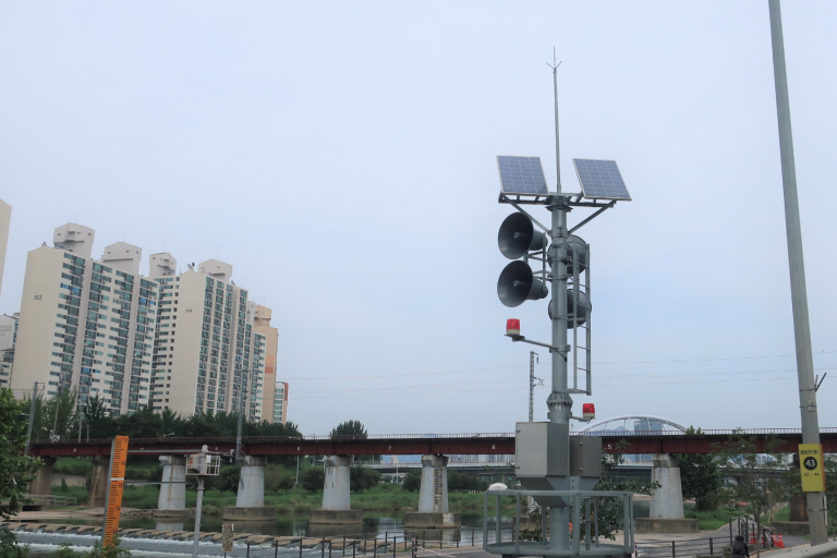 A tall pole with solar panels and loudspeakers stands by a river, near a railway bridge and high-rise buildings under a cloudy sky.