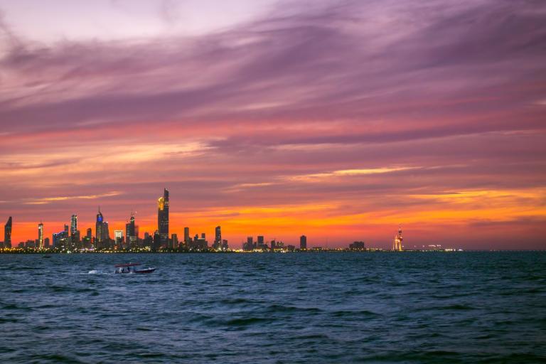 City skyline at sunset with colorful clouds above, a large body of water in the foreground, and a small boat on the water.
