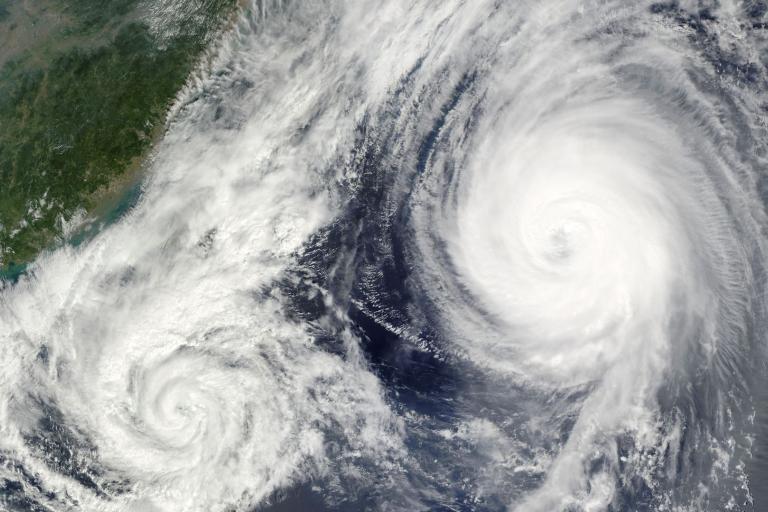 Satellite image of two large cyclones in the ocean near a landmass, with swirling cloud patterns visible.