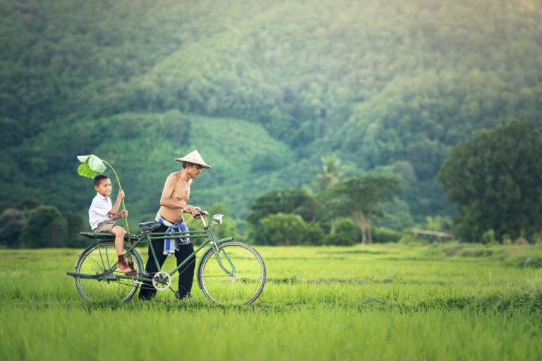 A shirtless man wearing a conical hat rides a bicycle through a lush green field, with a young boy holding a large leaf seated on the back.