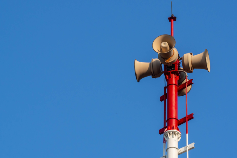 A red and white pole with several loudspeakers mounted on top, against a clear blue sky.