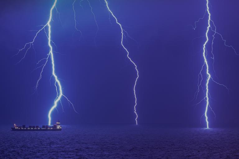 A ship sails on the ocean at night as two bright lightning bolts strike the water.
