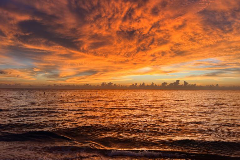 Sunset over the ocean with vibrant orange and yellow hues in the sky, reflecting on the water's surface. Waves gently lap against the shore under dramatic cloud formations.