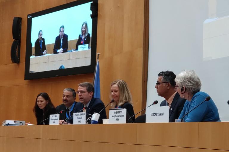 A group of people seated at a conference table with microphones in a meeting room. A screen displays their image. Plaques in front list their titles.