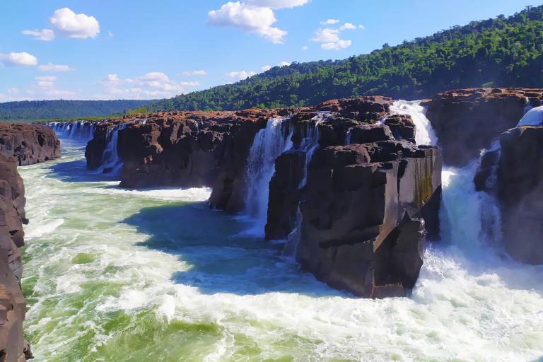 Waterfalls cascade over rocky cliffs into a turbulent river under a blue sky with scattered clouds, surrounded by a forested landscape.