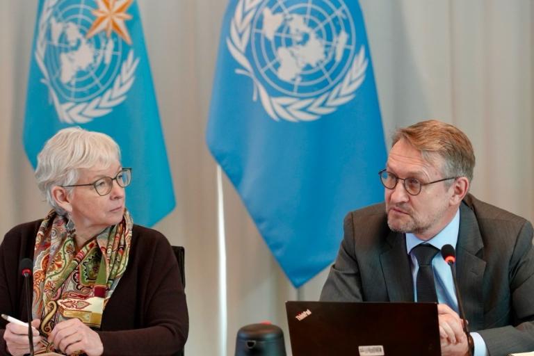 Two people sit at a table with microphones, engaged in discussion. Two United Nations flags are visible in the background.