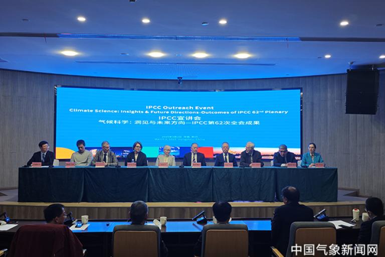 A panel of nine people seated at a table during an IPCC outreach event, with a large blue presentation screen behind them. Audience members are visible in the foreground.