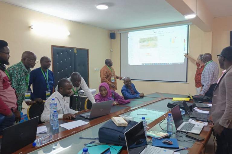 People engaged in a meeting, some seated at a long table with laptops, while one person presents information on a projected screen.