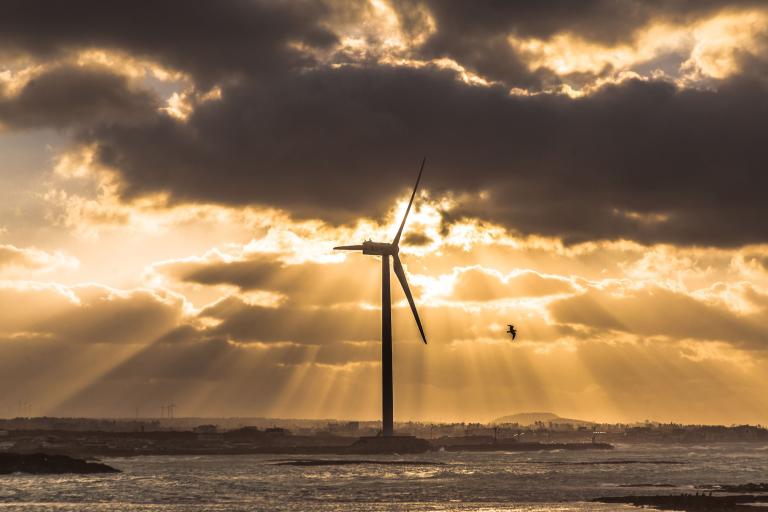 A wind turbine stands by the water at sunset, with clouds and sun rays in the sky and a bird flying nearby.