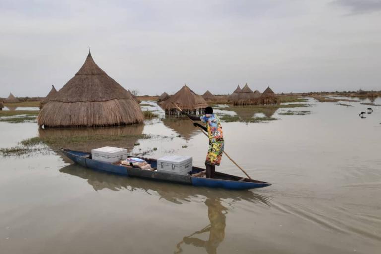 A person paddles a small canoe on a floodplain, passing traditional thatched huts partially submerged in water.