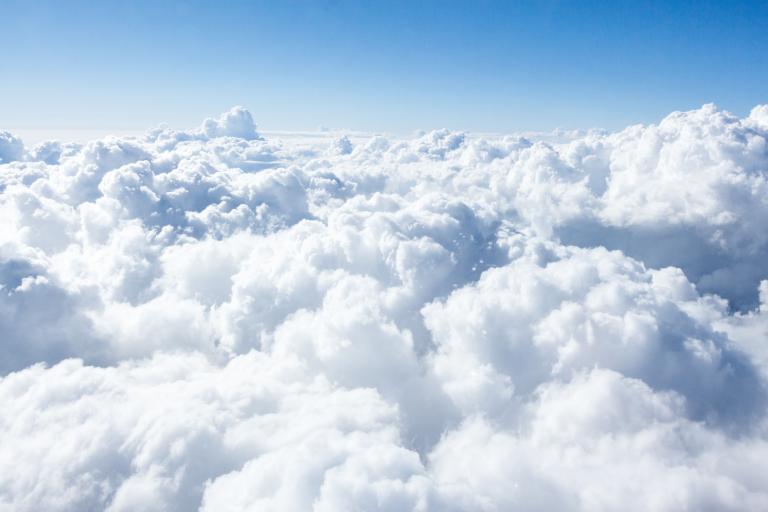 Fluffy white clouds fill the sky under a clear blue background, creating a soft, airy landscape from an aerial perspective.