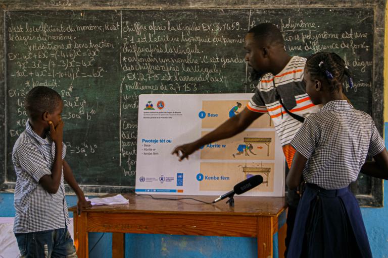 Teacher explaining a safety poster to two students in front of a chalkboard in a classroom. The chalkboard is filled with text, and a microphone is visible on the table.
