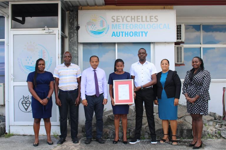 Seven people stand in front of the Seychelles Meteorological Authority building. One person holds a framed certificate.