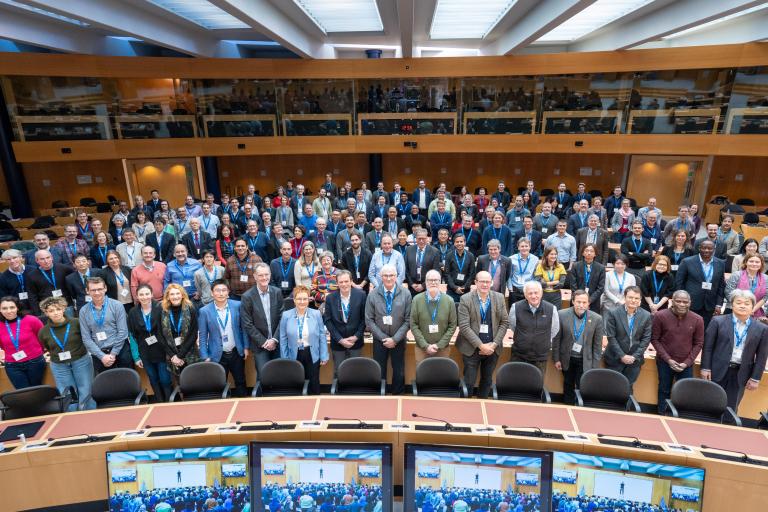 A large group of people pose for a group photo in a modern conference room with curved desks and computer monitors at the front.