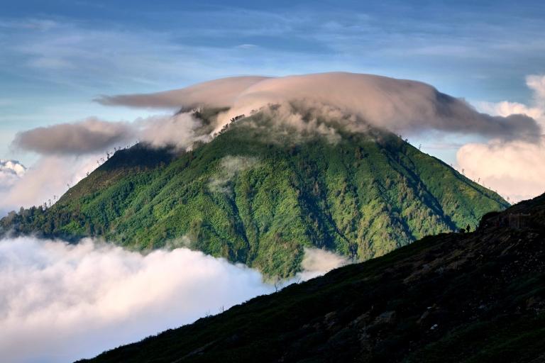 A green mountain peak is partially covered by a cap of clouds, with additional clouds surrounding its base under a clear blue sky.