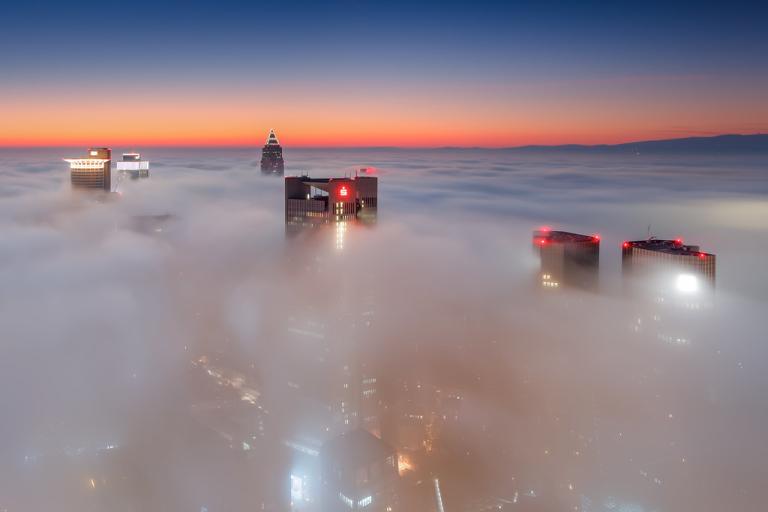 Tall city buildings rise above dense fog at sunrise, with the sky displaying a gradient from deep blue to orange on the horizon.