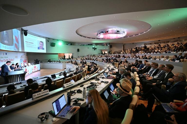A large conference room filled with delegates seated in rows, attending a formal panel discussion with presentations displayed on large screens.