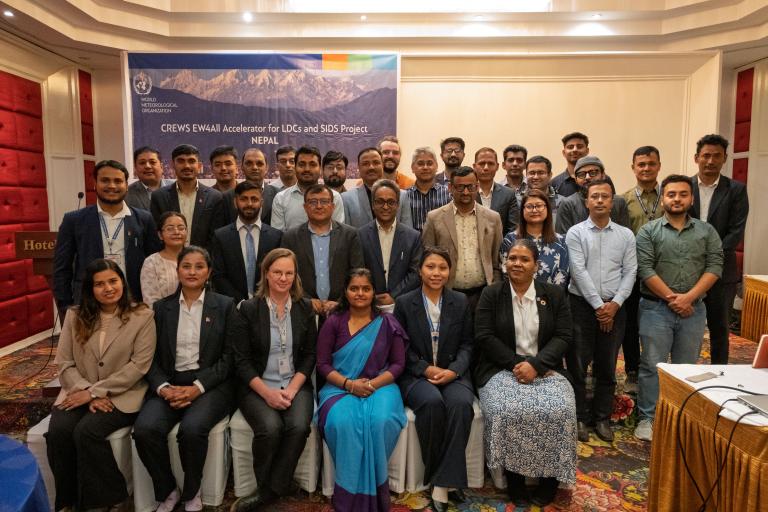 A group of men and women in business attire pose for a photo in a conference room with a banner reading "CREWS EW4All Accelerator for LDCs and SIDS Project Nepal" in the background.