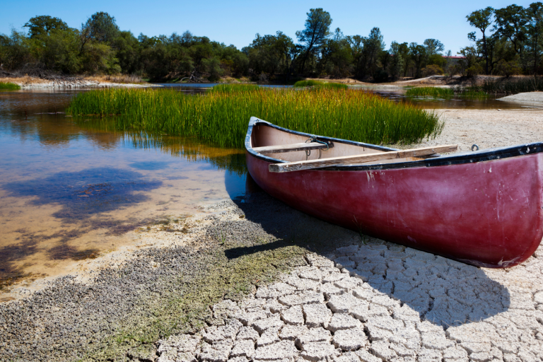A red canoe sits on cracked, dry ground beside a shrinking body of water with sparse vegetation and trees in the background.