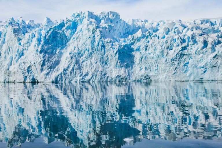 A glacier with jagged blue and white ice is reflected in calm, clear water under a partly cloudy sky.