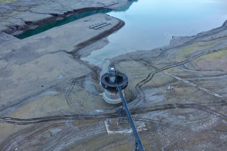 Aerial view of a partially dried reservoir with exposed land, visible tire tracks, and a circular water intake structure connected by a walkway.