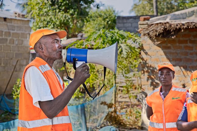 A man in an orange vest and cap speaks into a megaphone outdoors while another person in similar attire stands nearby.