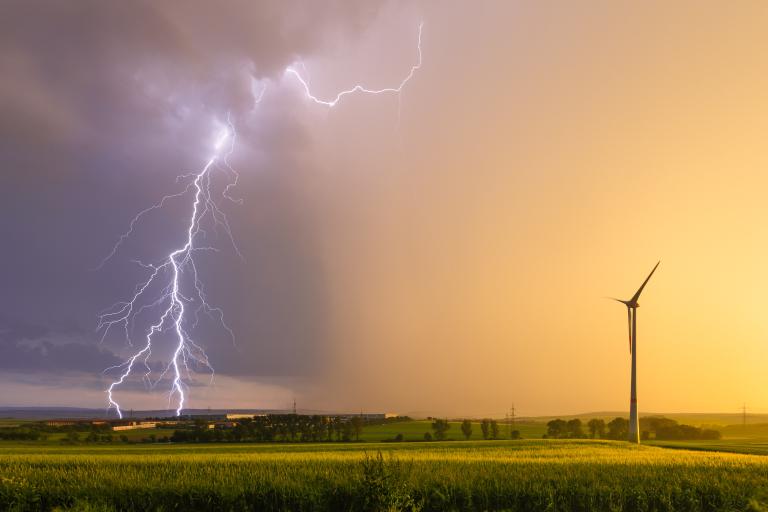 A lightning bolt strikes in the distance over fields at sunset, with a single wind turbine standing in the foreground.