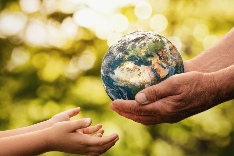 Two adult hands hold a small globe while two child hands reach out towards it, set against a blurred, green outdoor background.