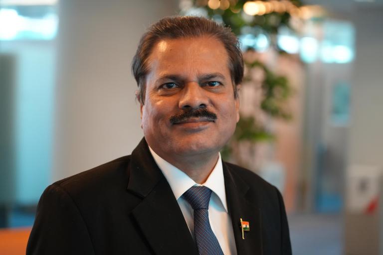 A middle-aged man in a suit and tie with a small Indian flag pin, standing indoors with blurred office background.