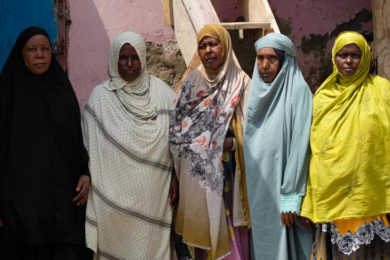Five women in colorful hijabs stand side by side in front of a pink wall and wooden stairs, looking at the camera.