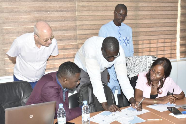 Five people in an office setting discuss documents spread out on a table, with laptops, water bottles, and papers visible.