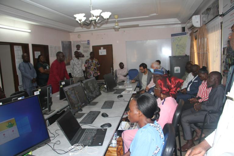 A group of people sits and stands around computer workstations in a classroom or office setting during a meeting or training session.