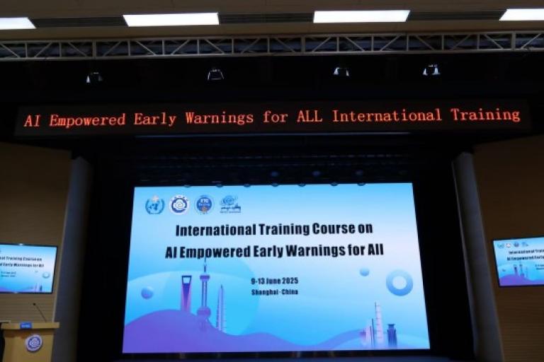 A conference stage displays a banner and digital sign for the "International Training Course on AI Empowered Early Warnings for All," held in Shanghai, China, June 2025.