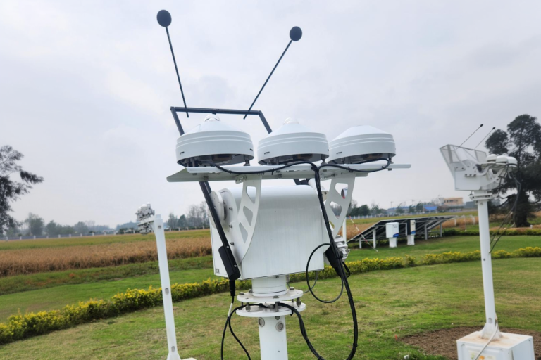 A weather monitoring instrument with three white sensor modules and antennae stands on a grassy field with crops and cloudy sky in the background.