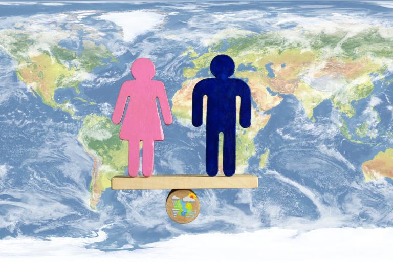 Pink female and blue male symbols stand balanced on a wooden plank over a globe, with a world map background, representing gender equality worldwide.